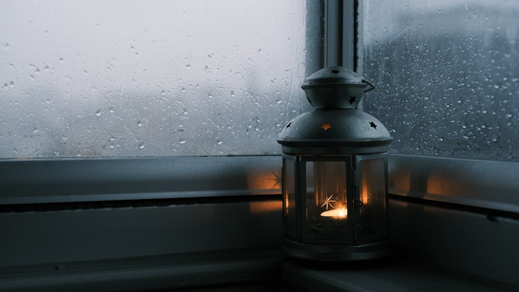 A flickering lantern by a rain-streaked window creates a cozy ambiance.